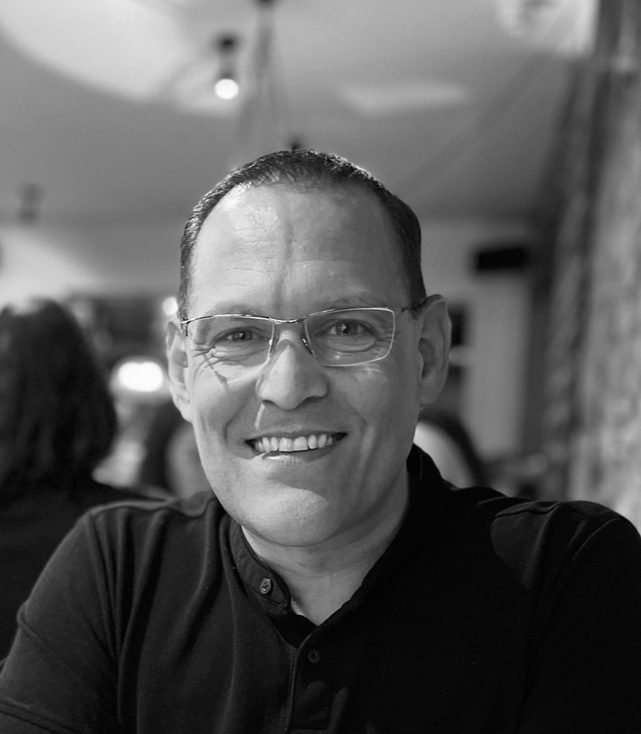 Ian Ashby Smiling man wearing glasses, seated in a casual setting with a blurred background.